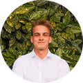 Young man in a white shirt smiling in front of green leafy background.